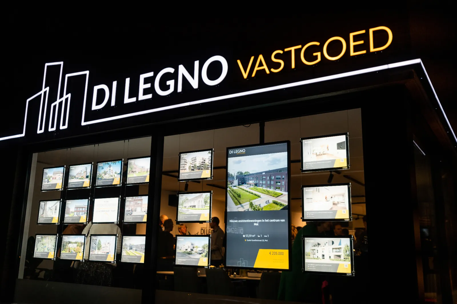 De verlichte etalage van Di Legno Vastgoed 's avonds, met een staand scherm tussen de panden