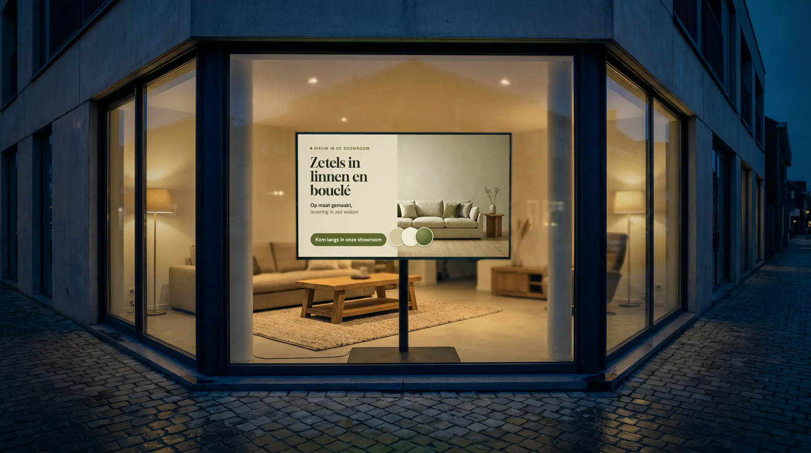 Hoekpand van een meubelzaak bij valavond, met een scherm op een voet in het raam voor een woonkamer met een zetel en een houten salontafel
